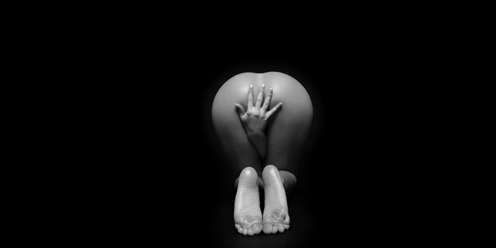 Erotischer Rückansicht einer knienden, nackten Frau, die ihre Hand zwischen den Beinen hindurch vor ihr Gesäß hält.
