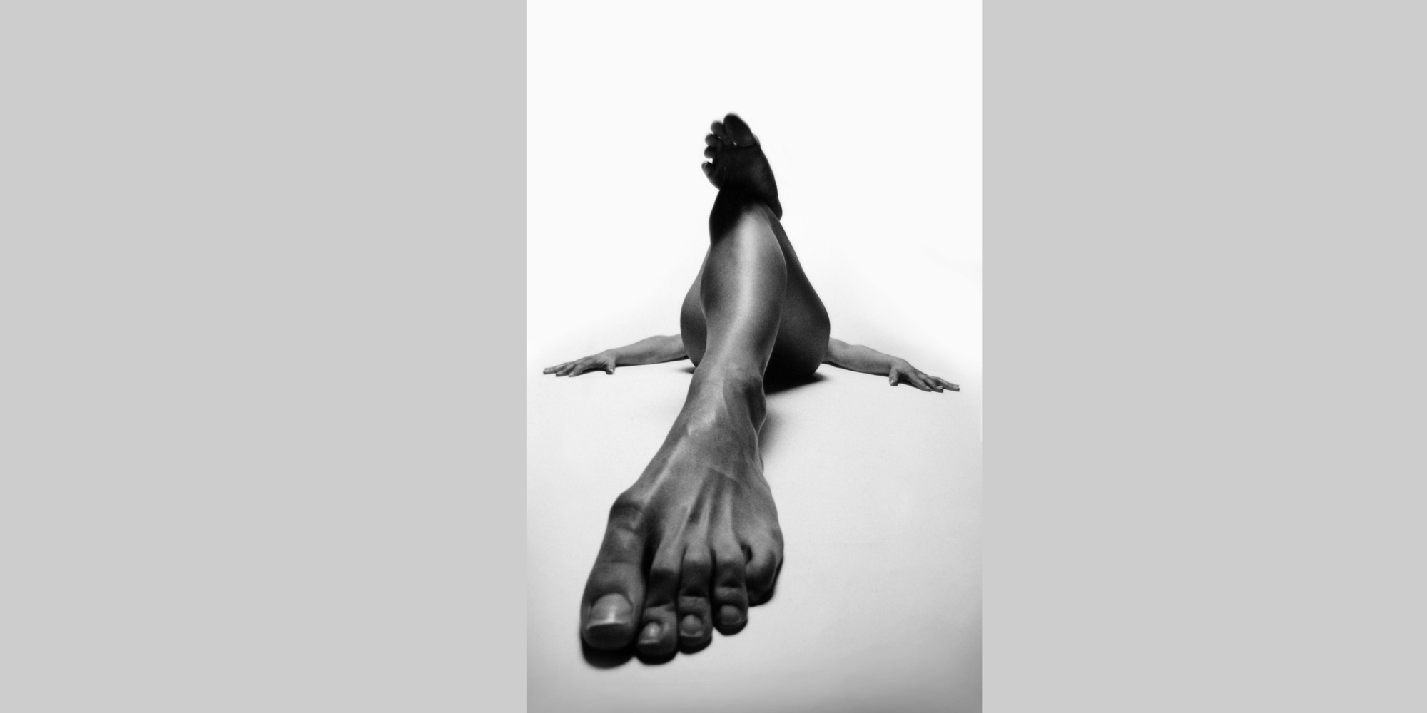 Frontale schwarz-weiße Ansicht einer liegenden, nackten Frau mit seitlich abgelegten Armen und ausgestrecktem Bein, wodurch ihr Fuß überdimensional groß wirkt, sie posiert für ästhetische erotische Aktfotografie.