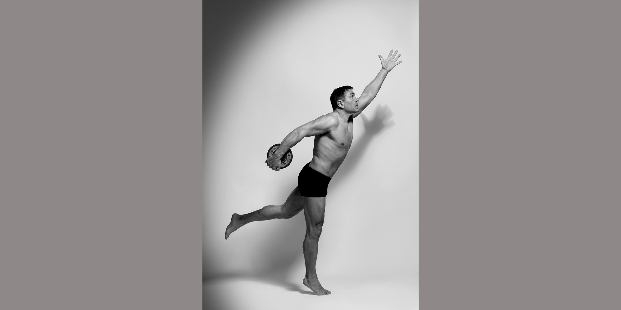 Seitliche Ganzkörperaufnahme eines sportlichen Mannes in Unterwäsche, der seine Arm und Bein diagonal ausstreckt und eine Hantel-scheibe festhält.