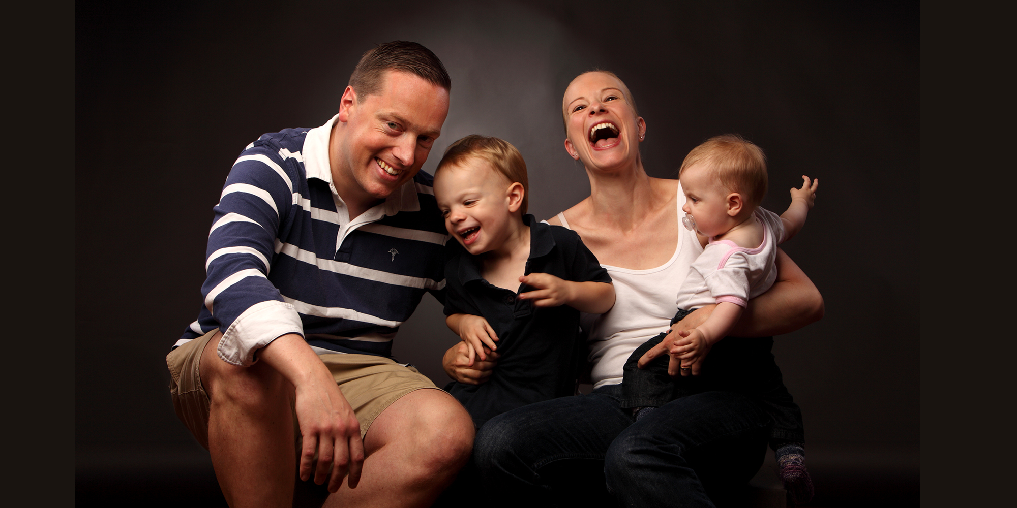 Portrait von einer Frau, die lauthals lacht und einen Säugling mit Schnuller auf dem Arm trägt; daneben sitzt ein Mann mit freundlichem Gesichtsausdruck und beide halten einen kleinen Jungen zwischen sich fest (Familienportrait).